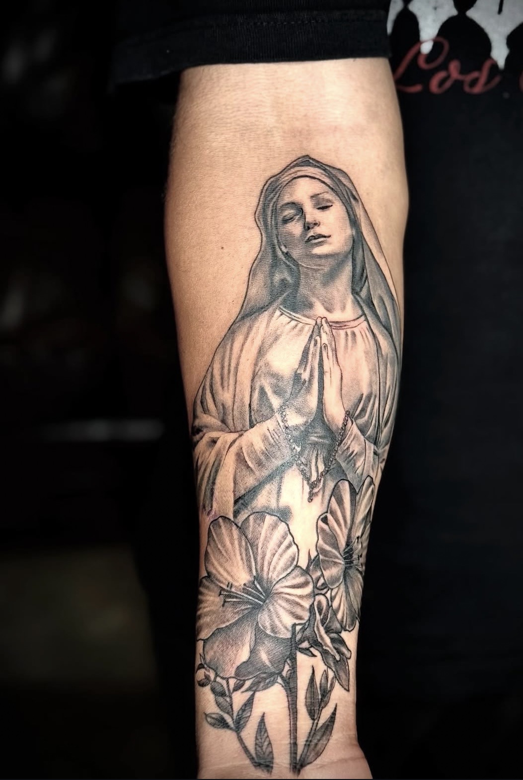 Black and grey tattoo of the Virgin Mary in prayer above lilies, by Aspired Ink